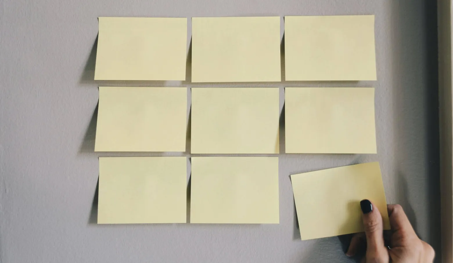 Leere Haftnotizen im Raster an einer Wand, eine Hand klebt den letzten Zettel an