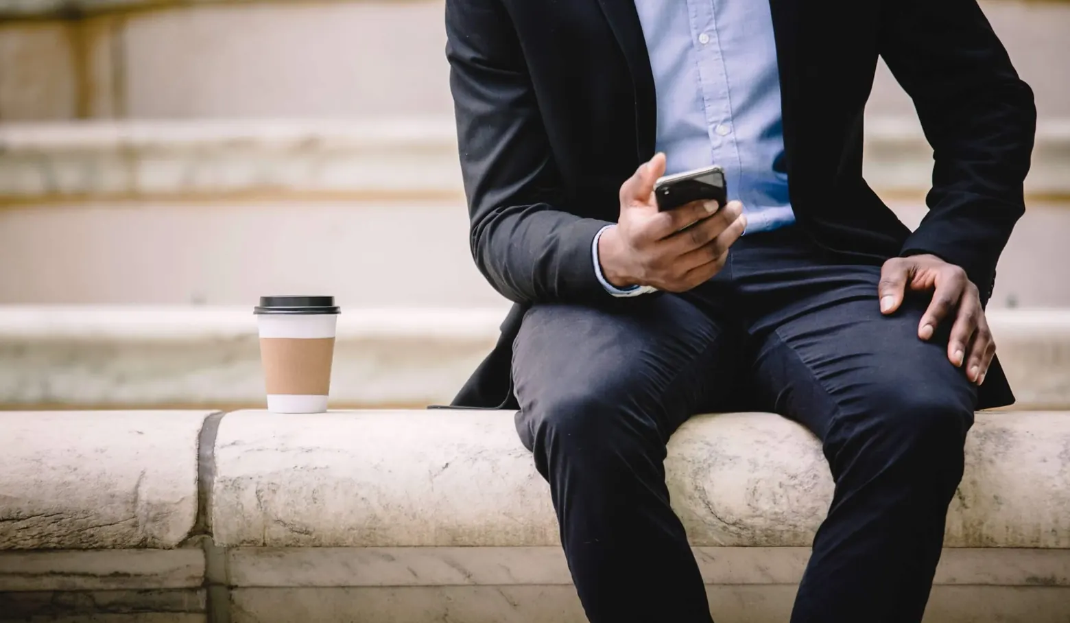 Mann im Anzug sitzt auf einer Steinbank und schaut auf sein Smartphone, daneben ein Kaffeebecher